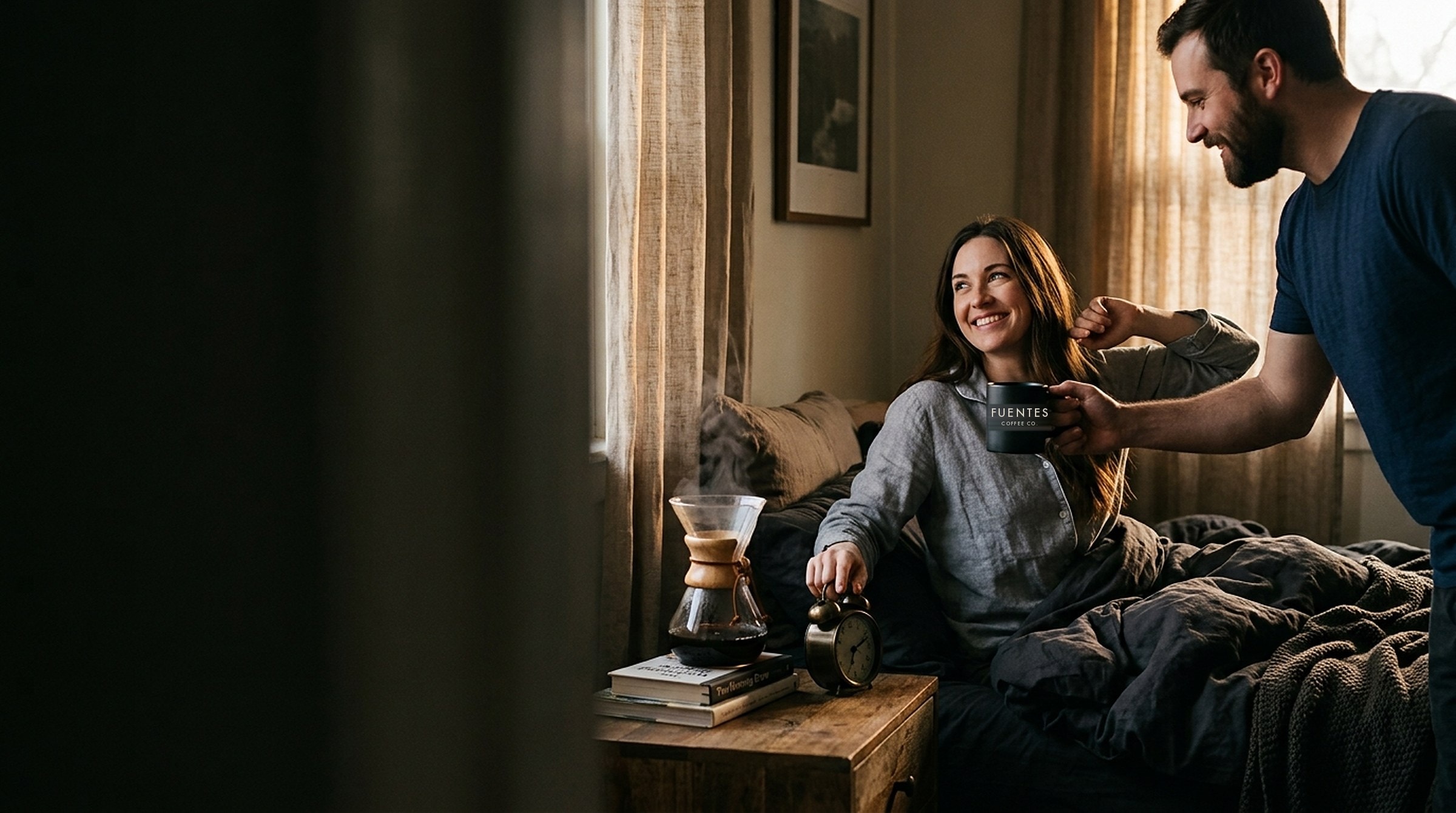 A woman waking up smiling in bed, switching off her alarm as her husband hands her a Fuentes Coffee mug, with a Chemex brewing on the nightstand